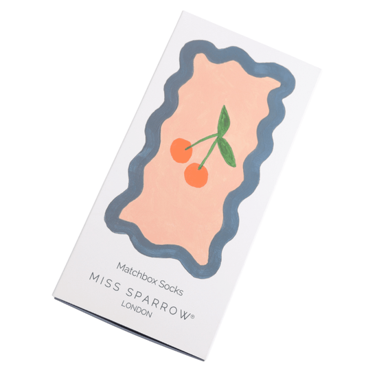 A white box with a cherry illustration in the middle on a pink background with blue border. Below are the workds matchbox socks - Miss Sparrow, London
