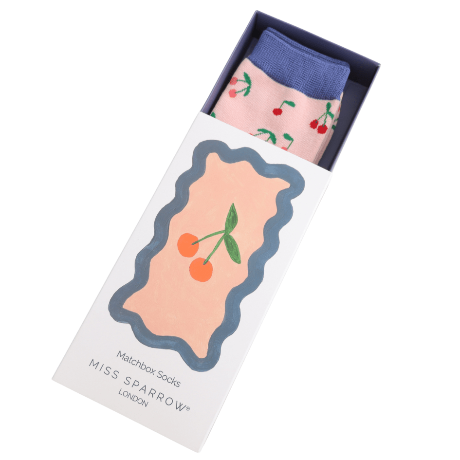 A matchbox style box half
 slid open, revealing the top of a pink pair of socks with blue cuff and cherry design
