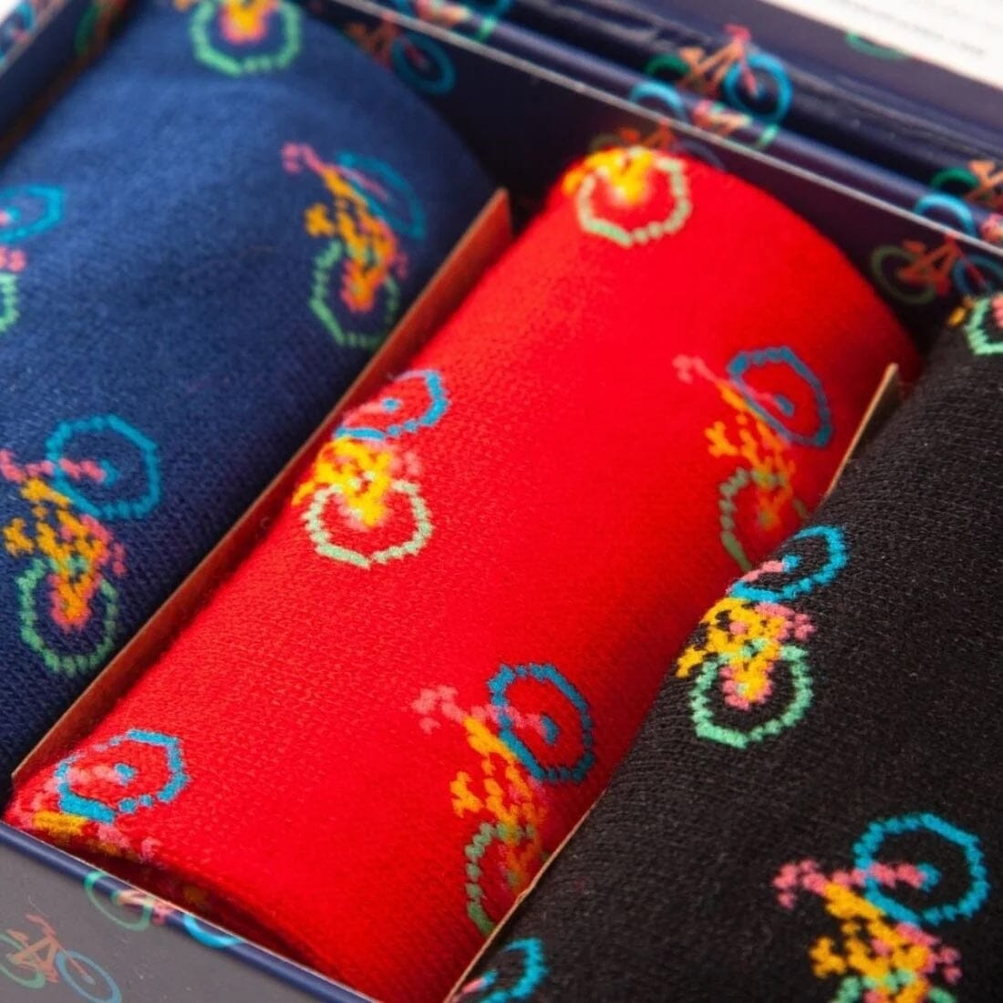 A close up of 3 pairs of bamboo socks ina box. One pair is blue, 1 red and 1 black, all have a epeated multi-coloured bicycle motif. 