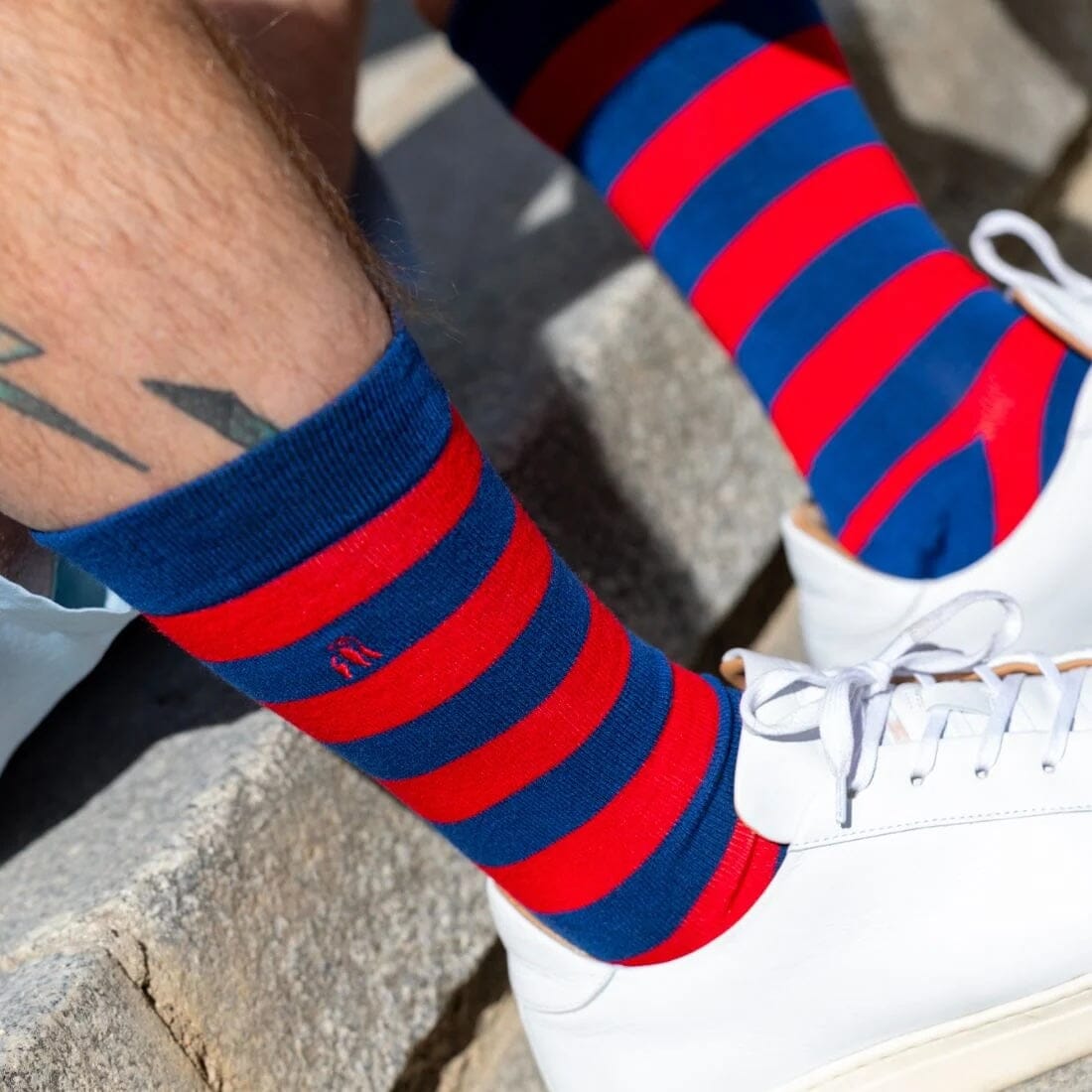 Red and blue striped socks worn with white sneakers on a stone surface.
