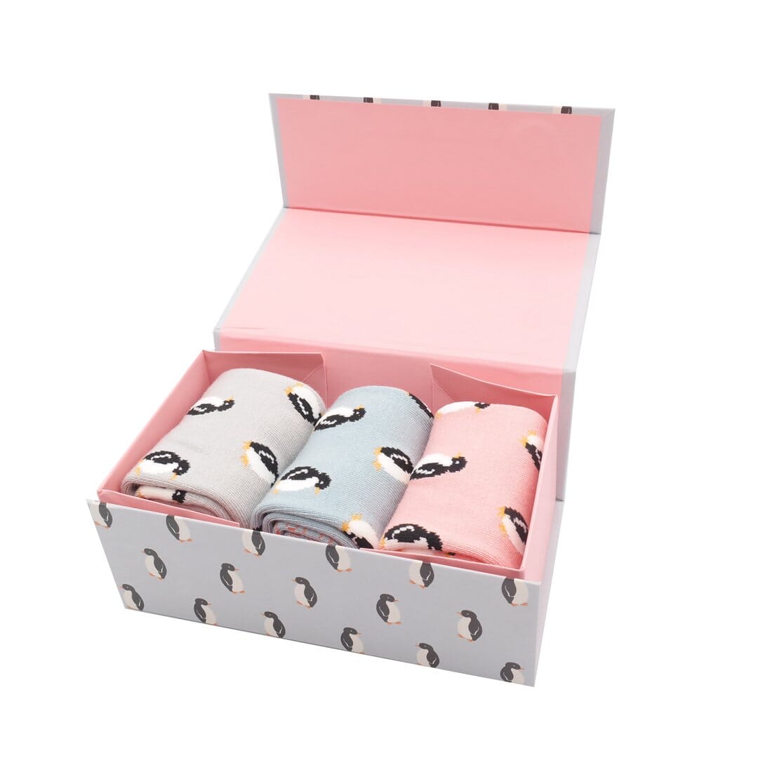 3 pairs of bamboo socks presented in a gift box.  The box is open.  1 pair of socks is pale grey, 1 pale blue, and one pale pink, and they all have penguins on them.  The box has the same penguin design. 
