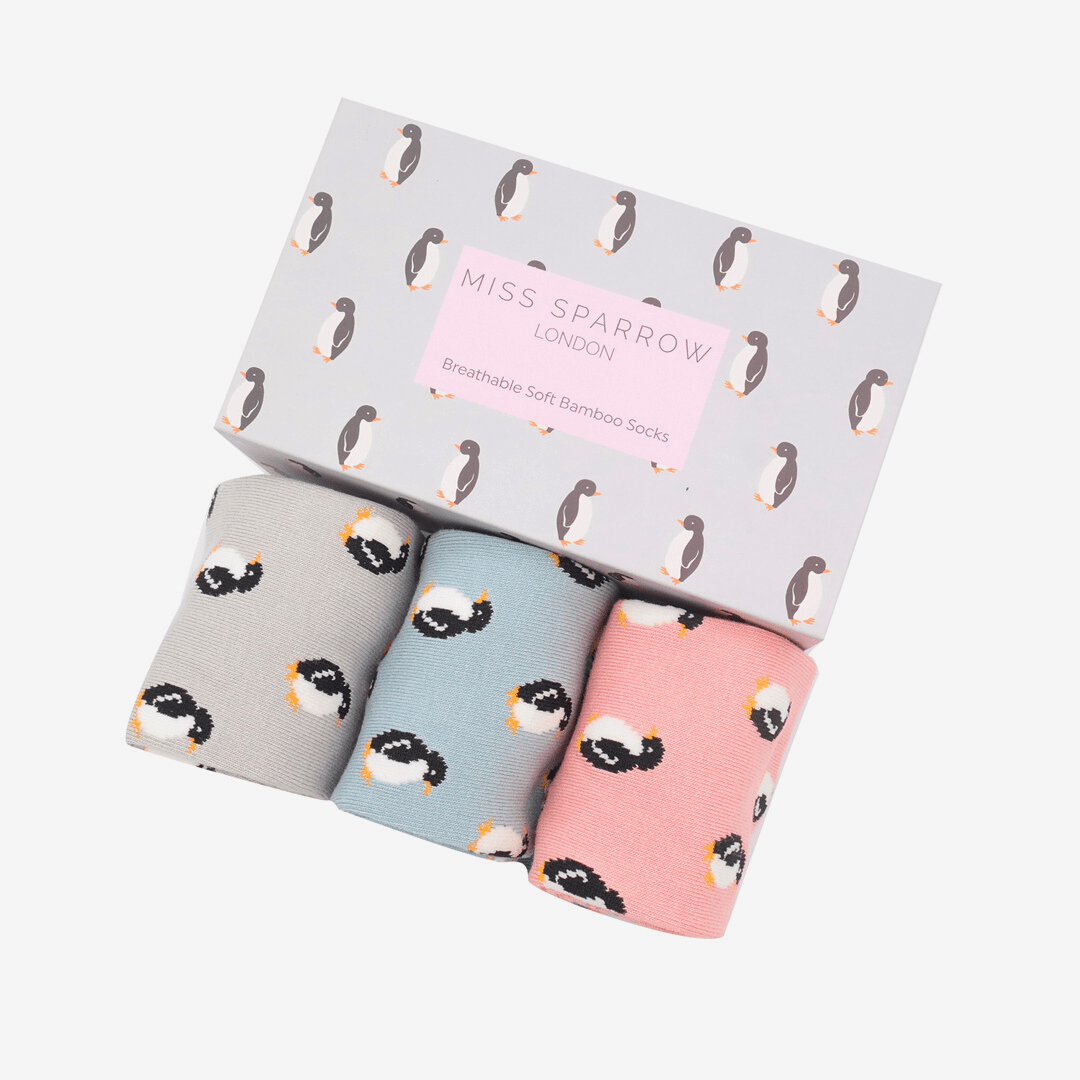 3 pairs of bamboo socks presented in a gift box.  The box is open.  1 pair of socks is pale grey, 1 pale blue, and one pale pink, and they all have penguins on them.  The box has the same penguin design. 