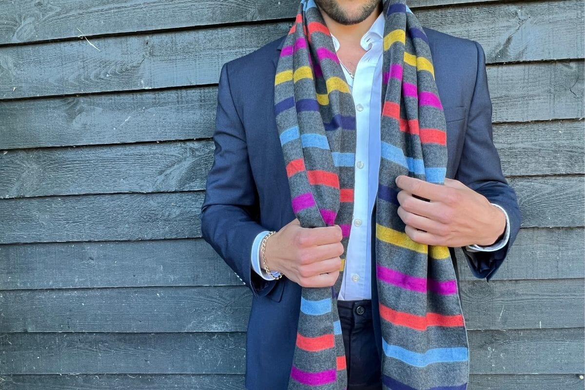 A man in a blue shirt and blue jacket wearing a scarf that is grey with colourful stripes - blue, purple, yellow, pink, orange.